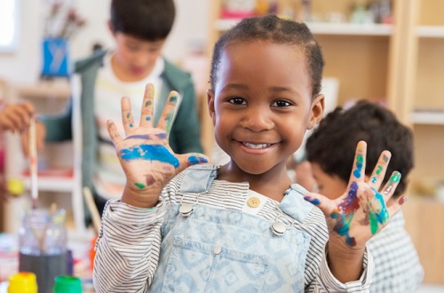 Young smiling child with painted hands in a classroom.